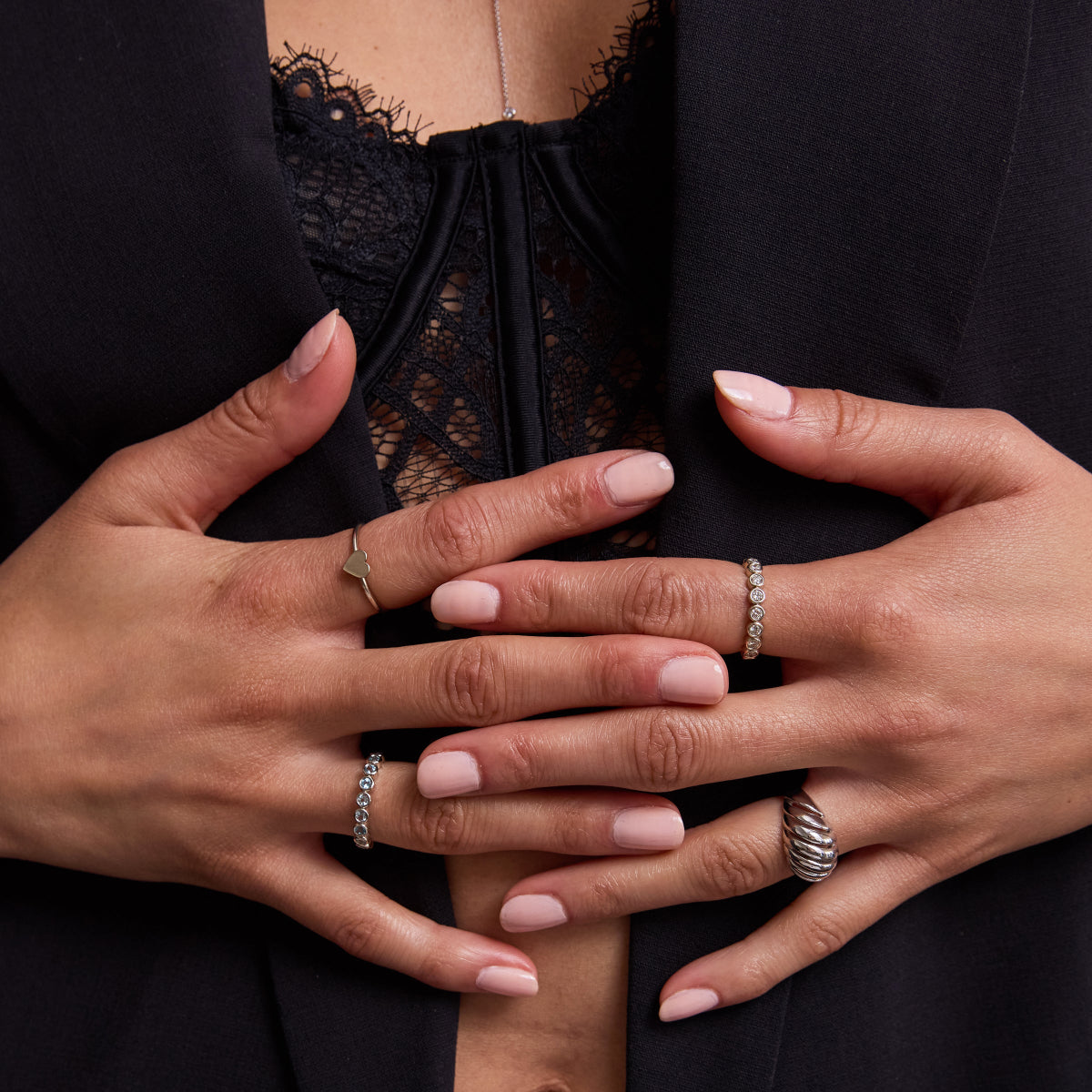A pair of hands wearing a white gold Heartfelt Ring, The Forget Me Not Bouquet Ring, The Evermore Ring and an Etched Paris Ring with light pink nails, styled with a lace black top and black blazer. 