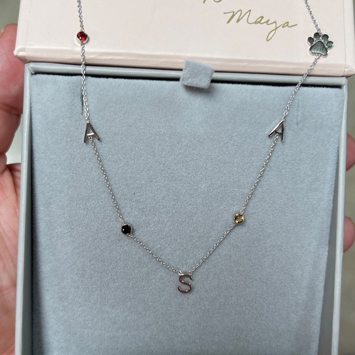 A Custom Gold Necklace in white gold with seven charms (two As, an S, three birthstones and a paw) in a Maya Brenner jewellery box. 