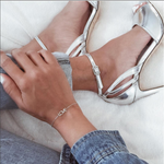 A woman wearing a Love Bracelet in white gold, resting her hand on her leg, wearing silver high-heeled sandals and denim jeans and top, against a neutral background.