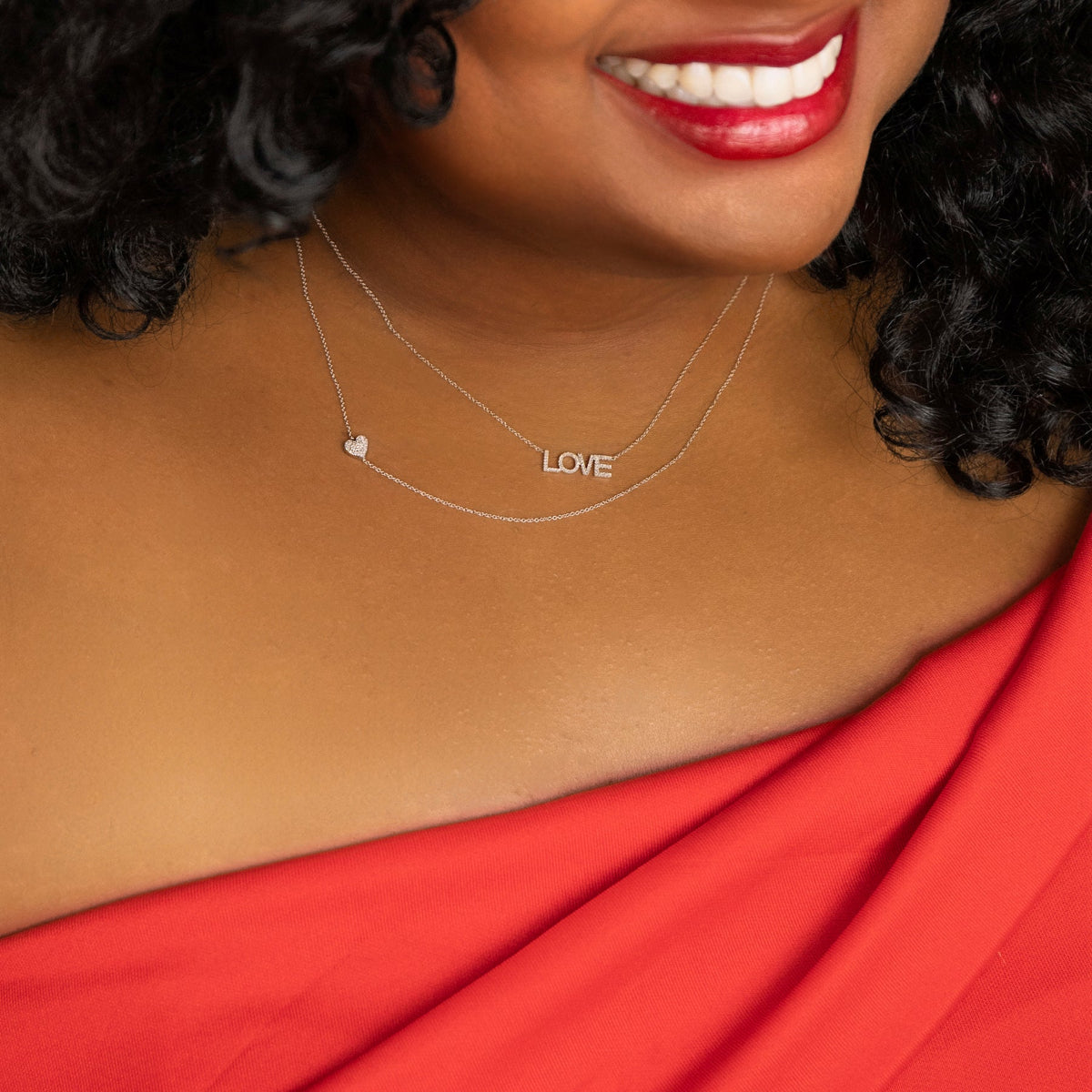A close-up of a woman's neck wearing a white gold 14k Gold Asymmetrical Charm Necklace - Pavé Heart and a Pavé LOVE Necklace, with black curly hair styled with a red top and lipstick. 