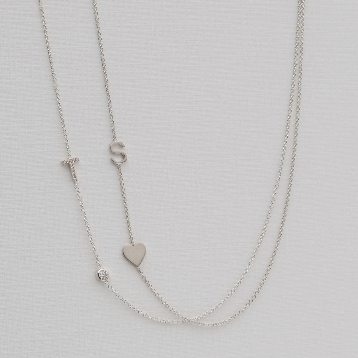A Monogram Necklace with Heart White Gold and a letter S charm, and a pavé Monogram Necklace with Diamond White Gold and a letter T charm, on a neutral background. 