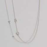 A Monogram Necklace with Heart White Gold and a letter S charm, and a pavé Monogram Necklace with Diamond White Gold and a letter T charm, on a neutral background. 