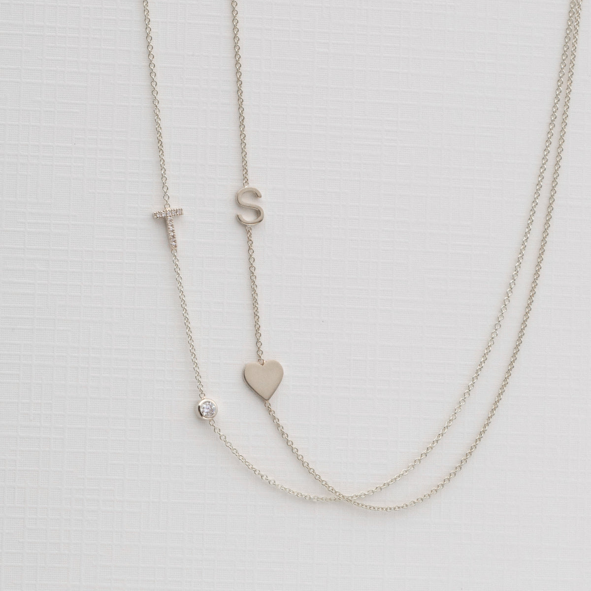 A Monogram Necklace with Heart White Gold and a letter S charm, and a pavé Monogram Necklace with Diamond White Gold and a letter T charm, on a neutral background. 