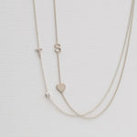 A Monogram Necklace with Heart White Gold and a letter S charm, and a pavé Monogram Necklace with Diamond White Gold and a letter T charm, on a neutral background. 