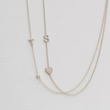 A Monogram Necklace with Heart White Gold and a letter S charm, and a pavé Monogram Necklace with Diamond White Gold and a letter T charm, on a neutral background. 