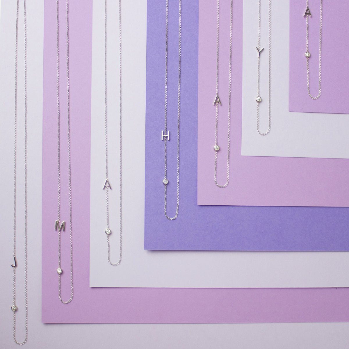 Seven Monogram Necklaces, each with a letter charm (J, M, A, H, A, Y and A) and a diamond charm, laid horizontally against variations of purple coloured paper.
