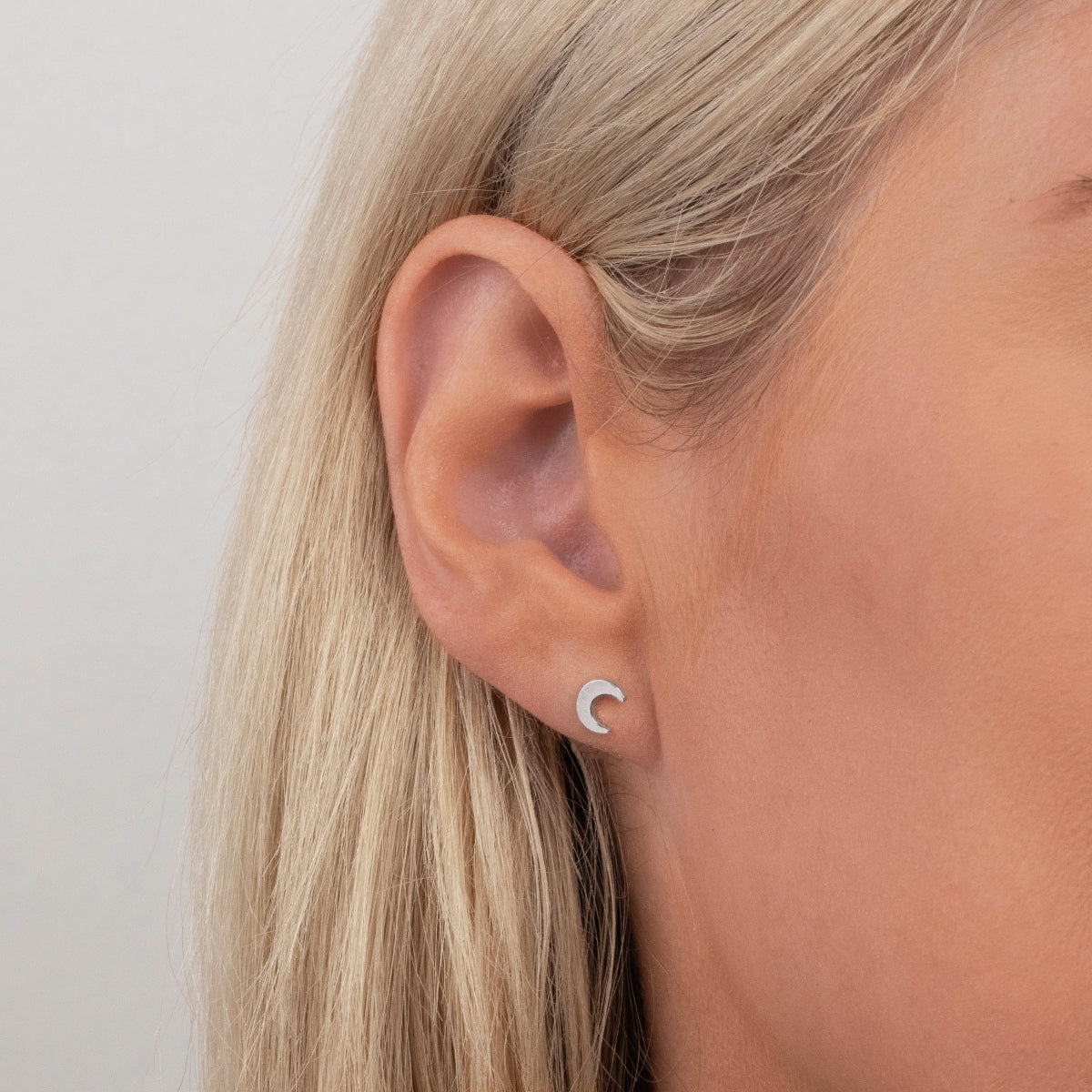 Close-up of a Charm Stud - Moon in white gold in a blonde woman's ear against a light grey background. 