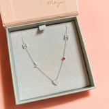 A Custom Gold Necklace in white gold with five charms (two pavé Hs, a pavé J, a pavé heart and a birthstone) in a jewellery box against a pink background.