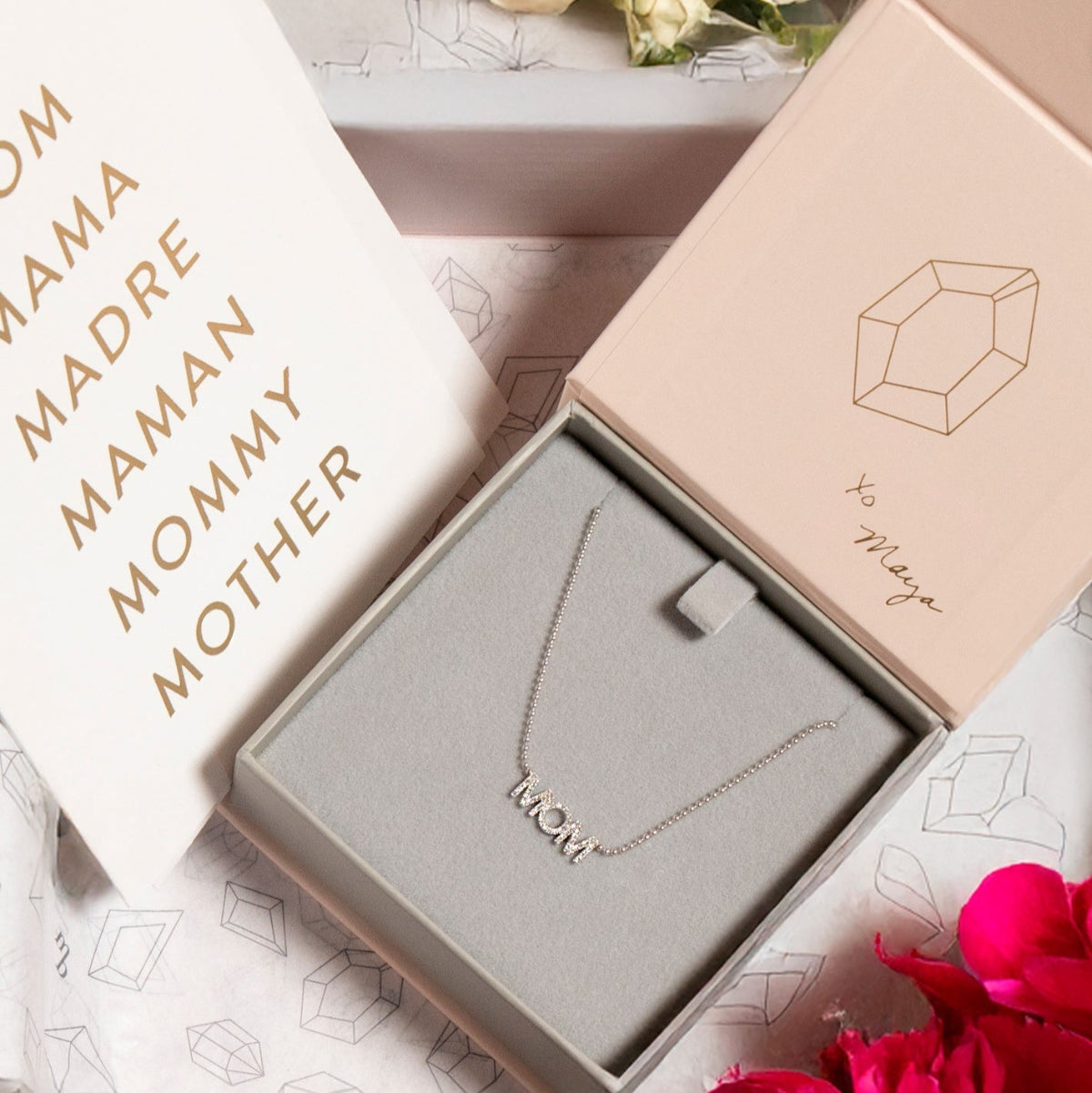 A Pavé MOM Necklace in white gold and a beaded chain displayed in a Maya Brenner jewellery box, accompanied by a paper featuring the words MOM, MAMA, MADRA, MAMAN, MOMMY, and MOTHER.