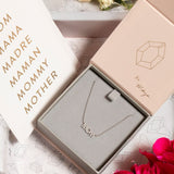 A Pavé MOM Necklace in white gold and a beaded chain displayed in a Maya Brenner jewellery box, accompanied by a paper featuring the words MOM, MAMA, MADRA, MAMAN, MOMMY, and MOTHER.