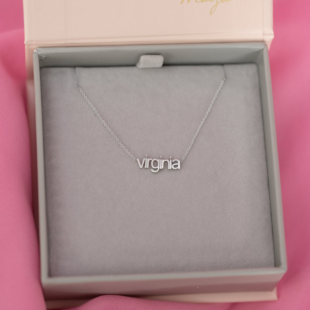 A white gold Petite Nameplate Necklace with 'virginia' in a Maya Brenner jewellery box with a pink background. 