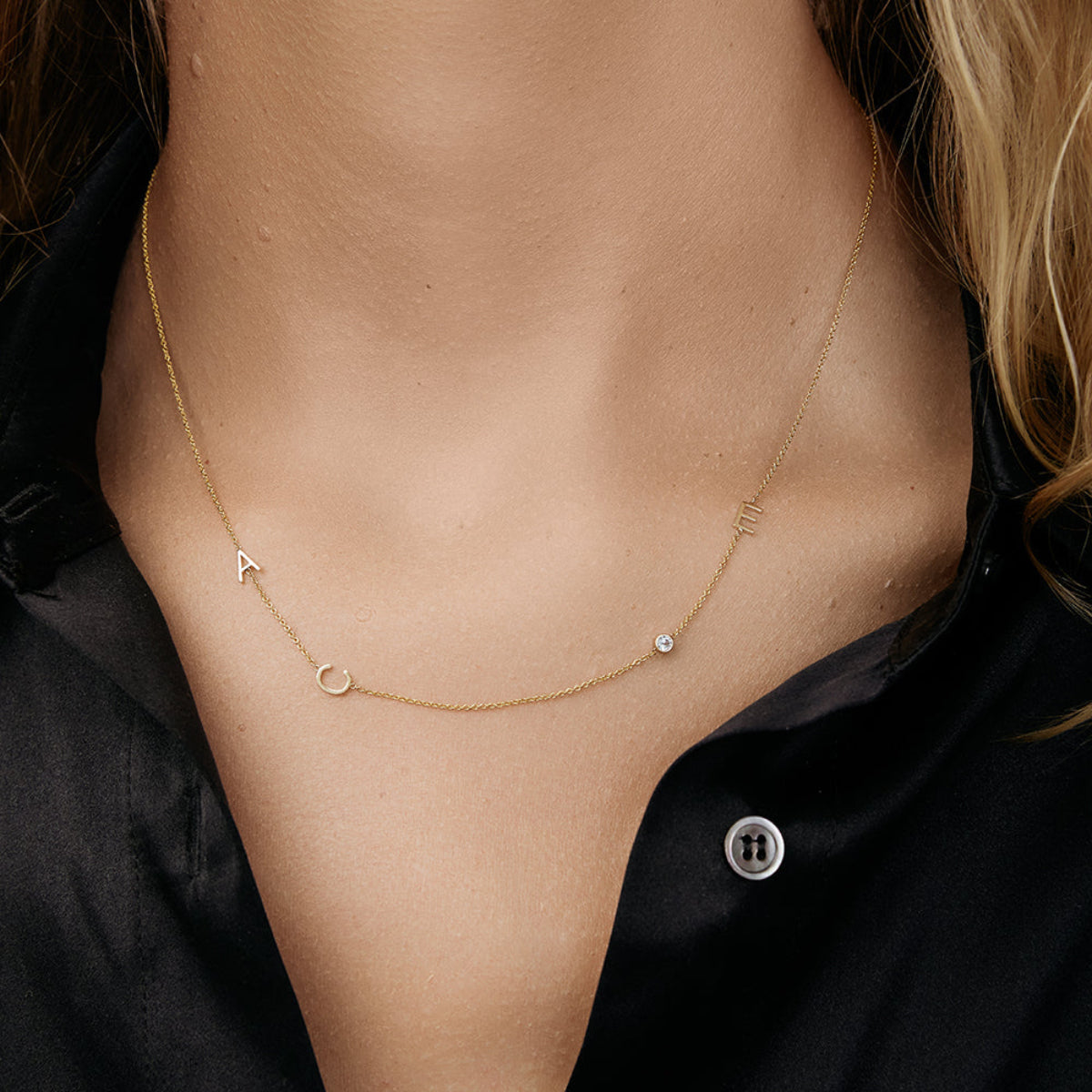 A close-up of a woman’s neck wearing a Custom Gold Necklace in yellow gold with the letters A and C on the left side and an E and birthstone on the right side, and a black shirt. 