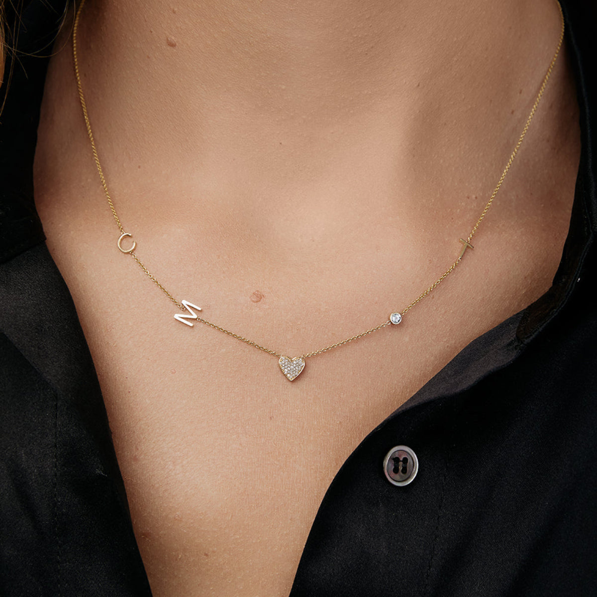 A close-up of a woman’s neck wearing a Custom Gold Necklace in yellow gold with five charms, three letters C, M, and T, a pavé heart and a birthstone, and a black shirt.