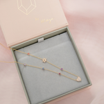 A Custom Gold Necklace in yellow gold with seven charms (five birthstones, a pavé Star of David and a pavé heart) in a Maya Brenner jewellery box against a light pink background.