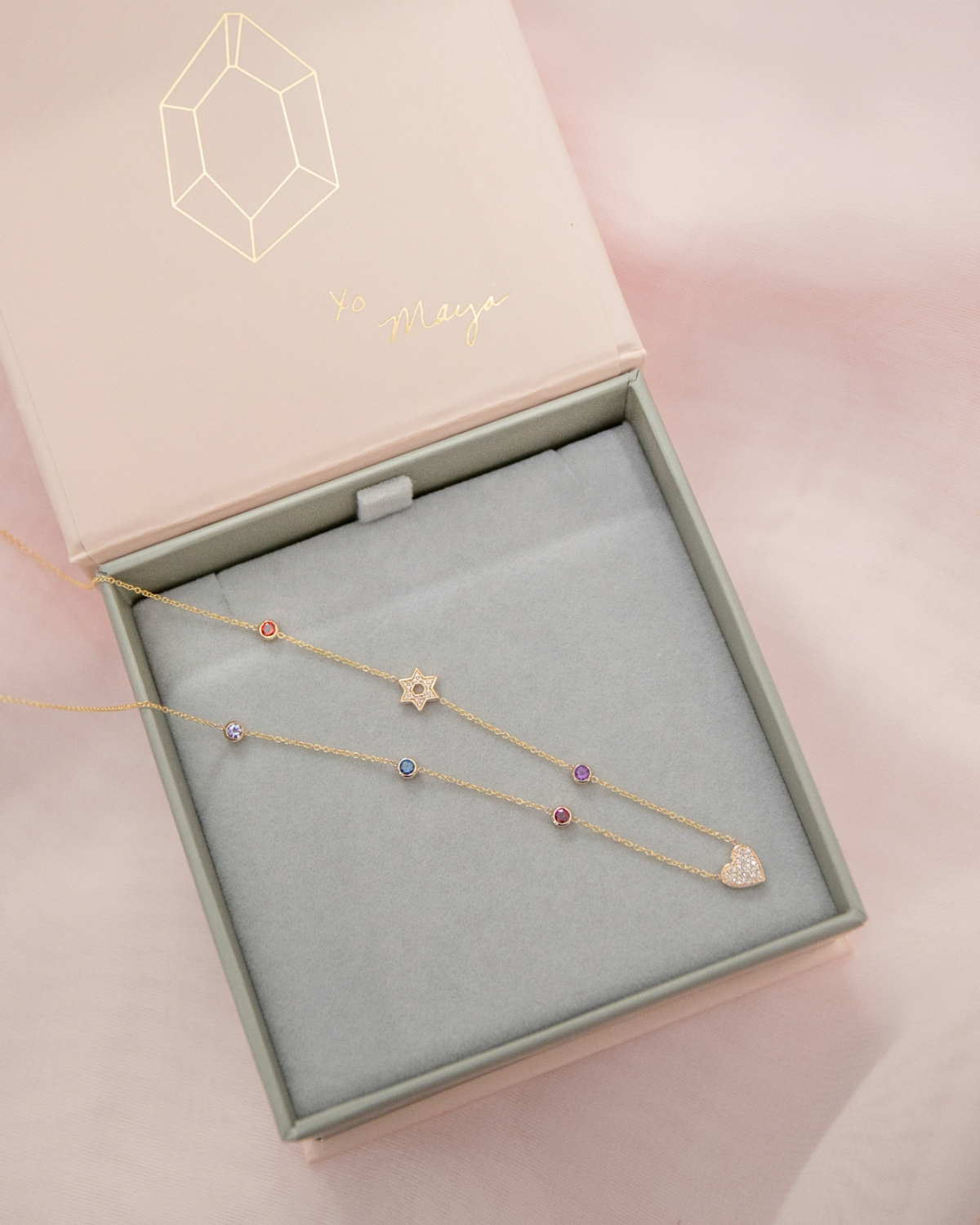 A Custom Gold Necklace in yellow gold with seven charms (five birthstones, a pavé Star of David and a pavé heart) in a Maya Brenner jewellery box against a light pink background.