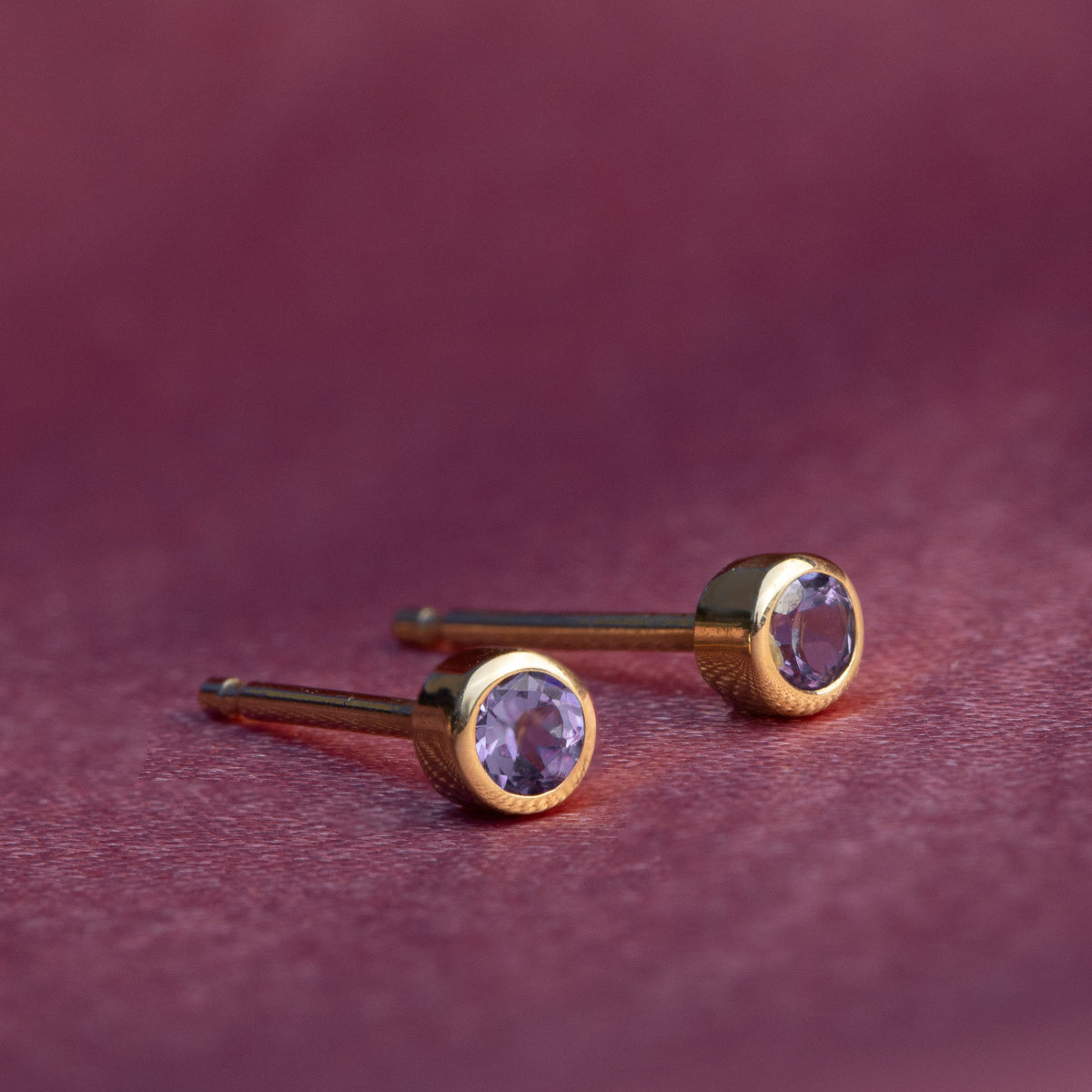 A pair of Birthstone Stud - Amethyst (February) in yellow gold against a dark purple background. 