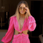 A blonde woman wearing a Zodiac Necklace - Aries, in a bright pink long sleeve dress and sunglasses against a dark background.