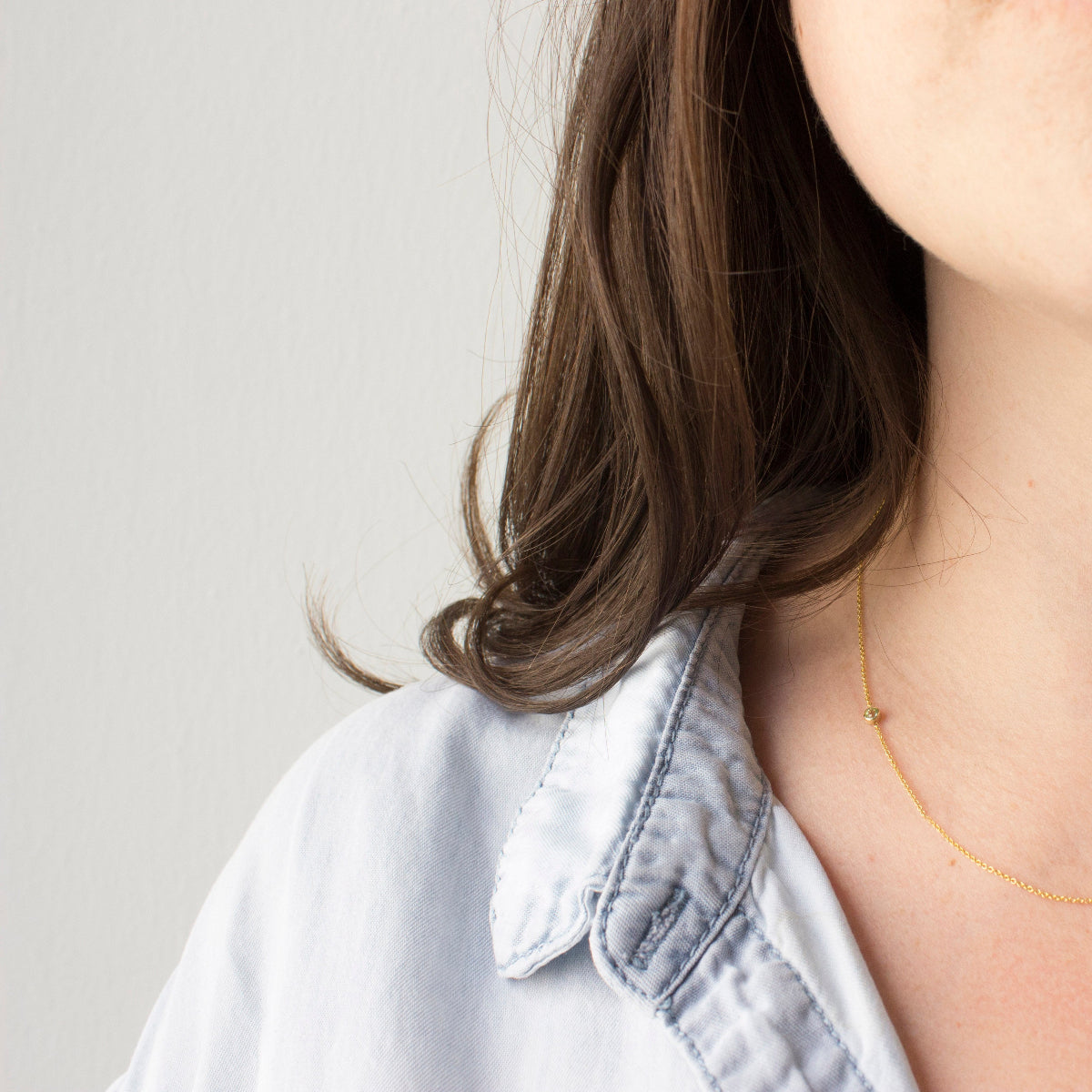 Close-up of a woman's neck and shoulder wearing a 14K Gold Asymmetrical Birthstone Necklace - Peridot (August) in yellow gold, with brown hair, styled in a light blue denim shirt against a light grey background.