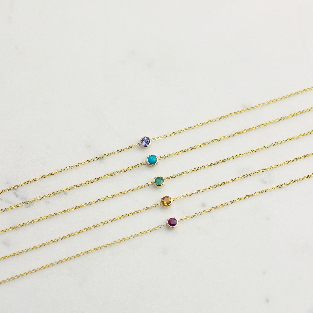 Five yellow gold 14K Gold Asymmetrical Birthstone Necklaces - Tanzanite (December), Turquoise (December), Emerald (May), Citrine (November) and Garnet (January) laid on a white background. 