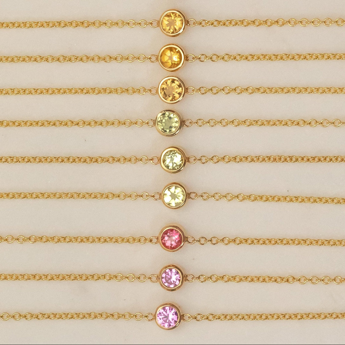 Twelve yellow gold 14K Gold Asymmetrical Birthstone Necklaces - three Diamond (April), three Citrine (November), three Peridot (August), one Garnet (January) and two Pink Tourmaline (October) laid against a neutral background. 