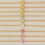 Twelve yellow gold 14K Gold Asymmetrical Birthstone Necklaces - three Diamond (April), three Citrine (November), three Peridot (August), one Garnet (January) and two Pink Tourmaline (October) laid against a neutral background. 