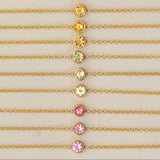 Twelve yellow gold 14K Gold Asymmetrical Birthstone Necklaces - three Diamond (April), three Citrine (November), three Peridot (August), one Garnet (January) and two Pink Tourmaline (October) laid against a neutral background. 