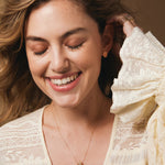 A woman wearing The Happiness Retreat Necklace, a Solid Eclipse Earring - Small and several rings, styled in a light yellow lace top with a warm background.