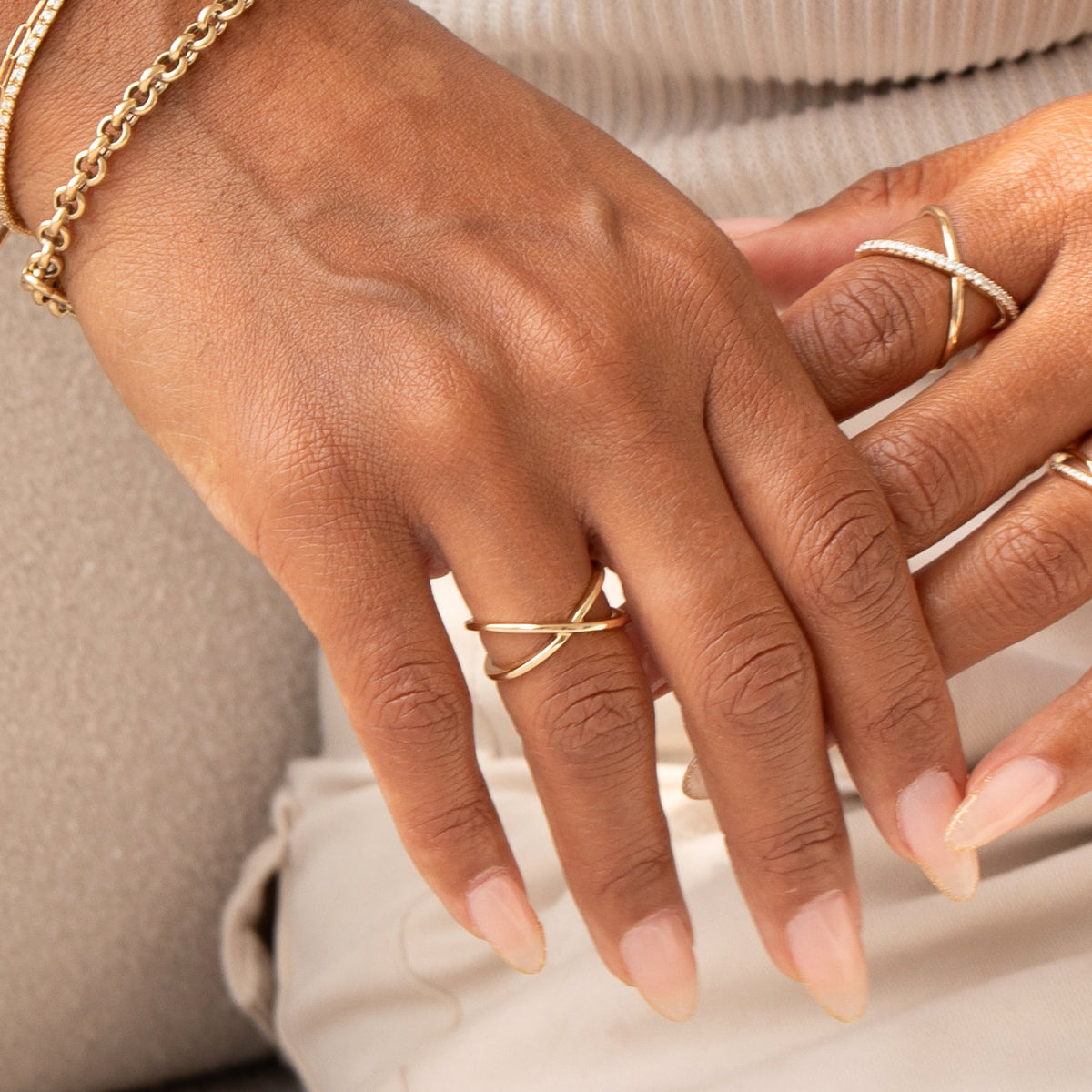 A pair of hands wearing a yellow gold Criss-Cross Ring, a Criss-Cross Diamond Daze Ring, a Rolo Bracelet and a Down the Line Bracelet, with light pink nails against a neutral background.