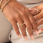 A pair of hands wearing a yellow gold Criss-Cross Ring, a Criss-Cross Diamond Daze Ring, a Rolo Bracelet and a Down the Line Bracelet, with light pink nails against a neutral background.
