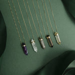 Five The Retreat Necklaces (Healing, Clarity, Protection, Courage and Happiness) laid against a dark green background.