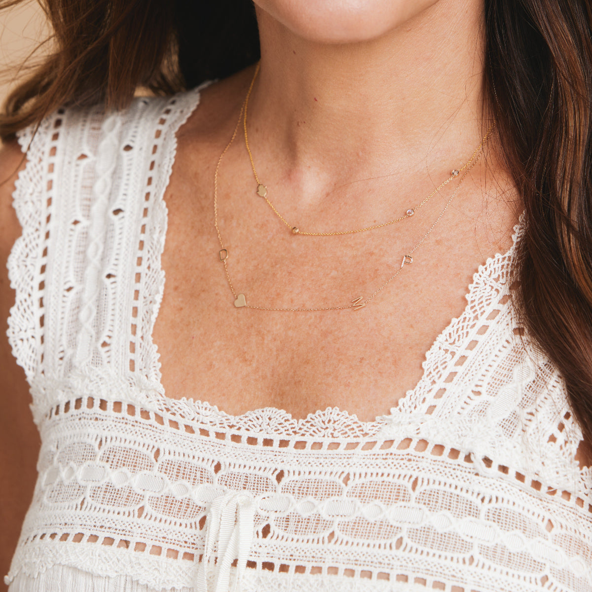 A close-up of a woman’s neck area wearing two Custom Gold Necklaces in yellow gold, one with three birthstones and a hand charm, and the other with letter charms D, P, and M and a heart, and a white lace top.