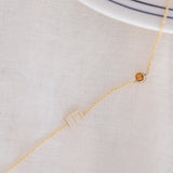 A Custom Gold Necklace in yellow gold with a letter E charm and a birthstone, on an off-white fabric background. 