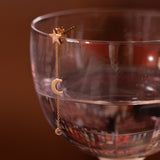 A Luna Spritz Earring on top of a cocktail glass against a brown background. 