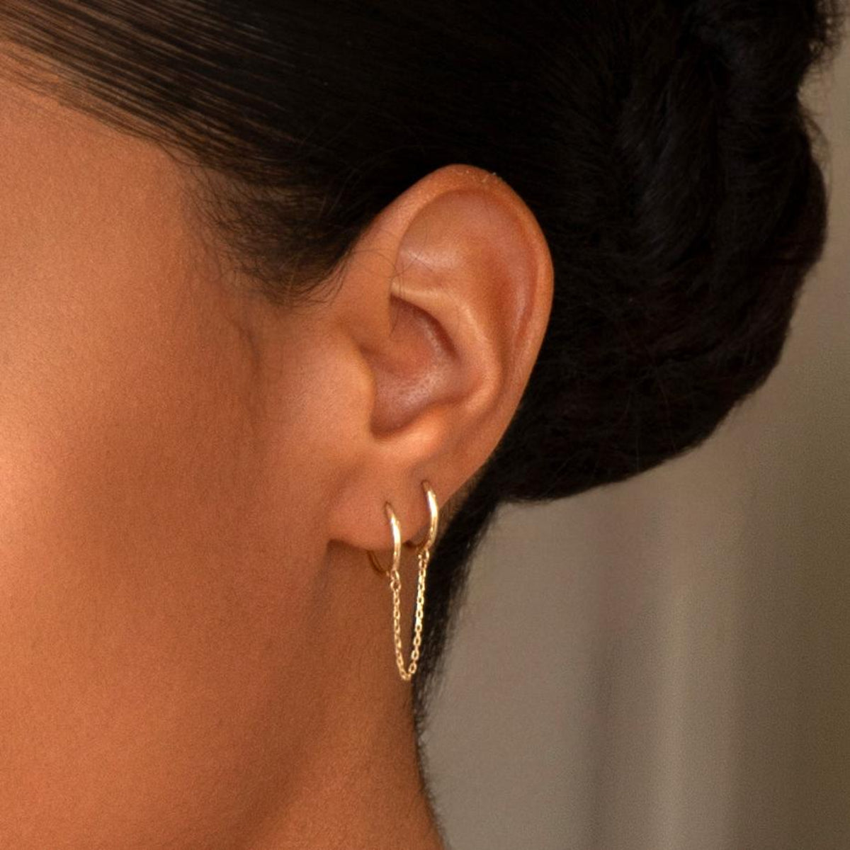 Close-up of a Double Huggie Earring with Chain being worn on woman's ear with black hair against a neutral background. 



