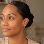 A woman wearing a Double Huggie Earring with Chain with a black bun looking to the side, against a blurred background. 