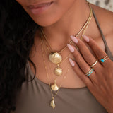 A close-up of a woman wearing a Chic Cuban Curb Chain with The Golden Venus Shell, an Element Long Link Chain with a Golden Scallop Shell, a Classic Bead Chain with a Golden Coast Shell, The Slide Chain with a Golden Spiral Shell, and several Bouquet Rings with a light brown singlet top. 
