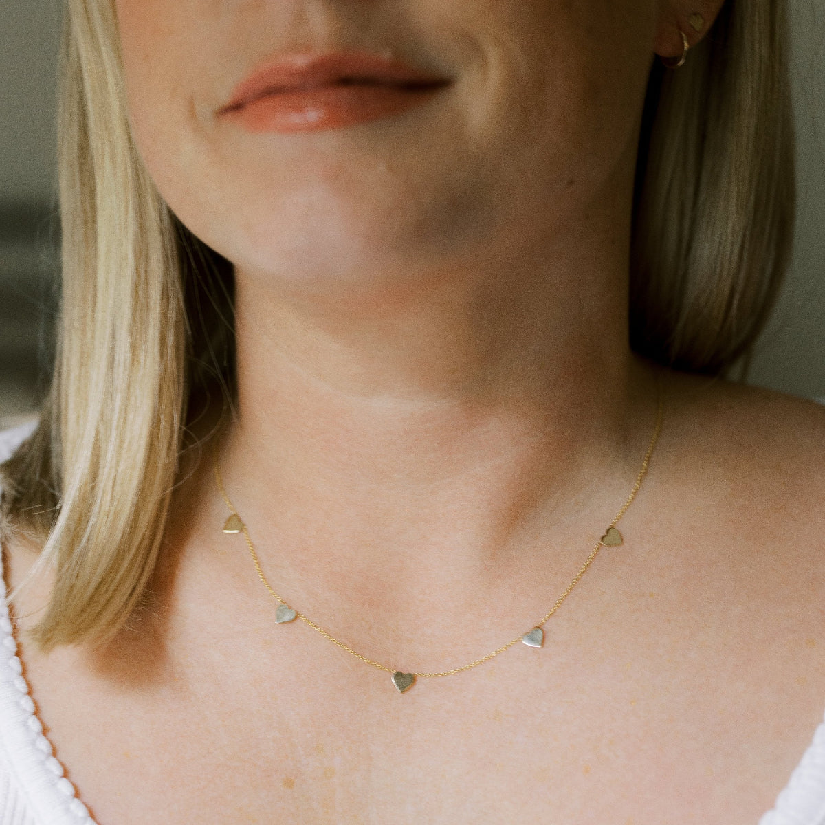Close up of a  woman's neck wearing a 5 of Hearts Necklace in yellow gold with a white top and her blonde hair partially visible. 