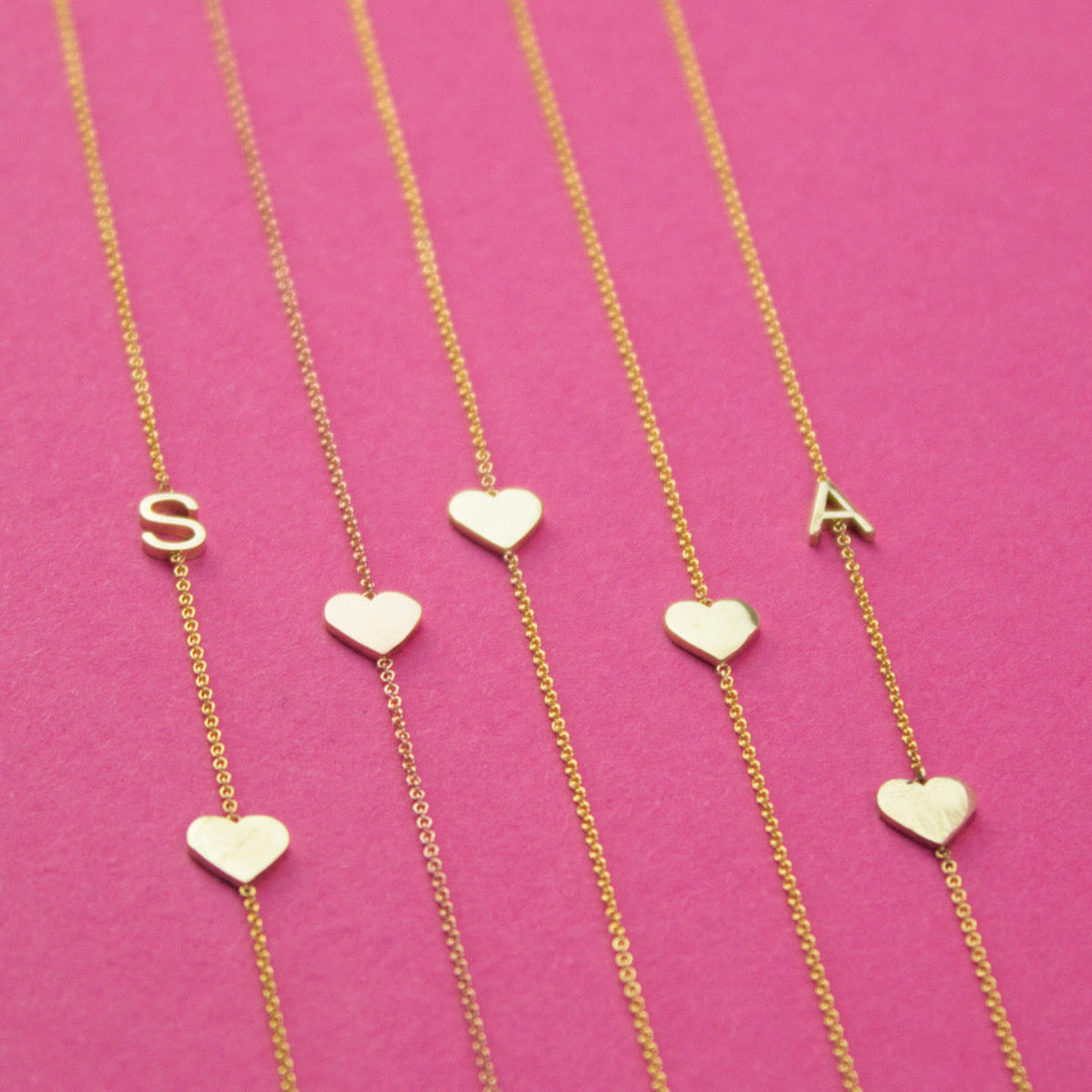 Five yellow gold necklaces laid out against a bright pink background, two Monogram Necklaces with Heart Yellow Gold with the letters S and A and three heart charm necklaces.