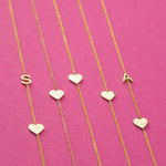 Five yellow gold necklaces laid out against a bright pink background, two Monogram Necklaces with Heart Yellow Gold with the letters S and A and three heart charm necklaces.