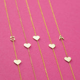 Five yellow gold necklaces laid out against a bright pink background, two Monogram Necklaces with Heart Yellow Gold with the letters S and A and three heart charm necklaces.