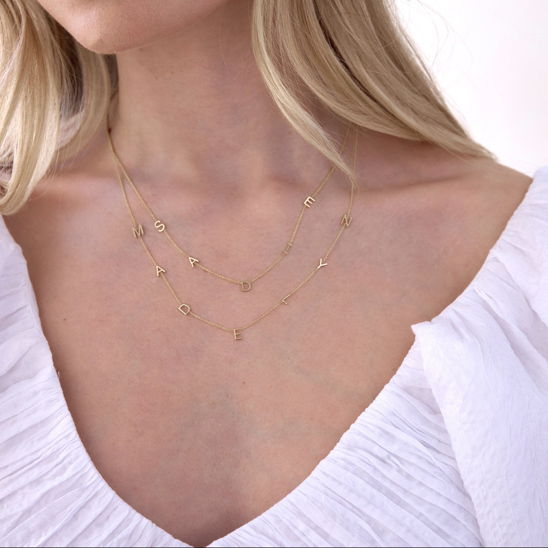 A close-up of a woman’s neck area wearing two Custom Gold Necklaces in yellow gold, one with five letter charms (S, A, D, I and E) and the other with seven letter charms (M, A, D, E, L, Y and N), with blonde hair and a white top against a white background.
