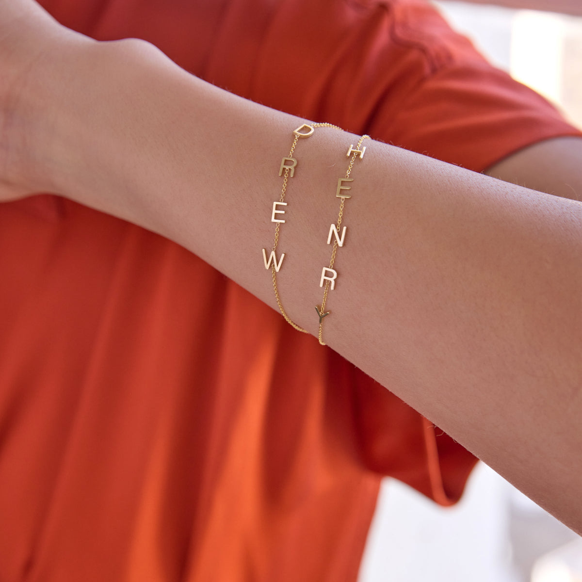 A close-up of an arm wearing yellow gold Custom Gold Bracelets, one with four letters and one with five letters, with a dark orange top.