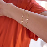 A close-up of an arm wearing yellow gold Custom Gold Bracelets, one with four letters and one with five letters, with a dark orange top.