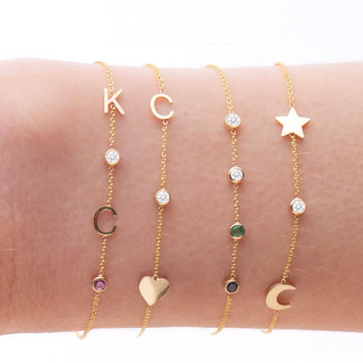 A wrist wearing four yellow gold Custom Gold Bracelets: two with three charms (C, a diamond birthstone and a heart, and a star, a diamond birthstone and a moon) and two with four charms (K, C, diamond and amethyst birthstones, and two diamonds, one emerald and one sapphire), on a white background.