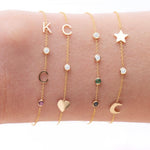 A wrist wearing four yellow gold Custom Gold Bracelets: two with three charms (C, a diamond birthstone and a heart, and a star, a diamond birthstone and a moon) and two with four charms (K, C, diamond and amethyst birthstones, and two diamonds, one emerald and one sapphire), on a white background.