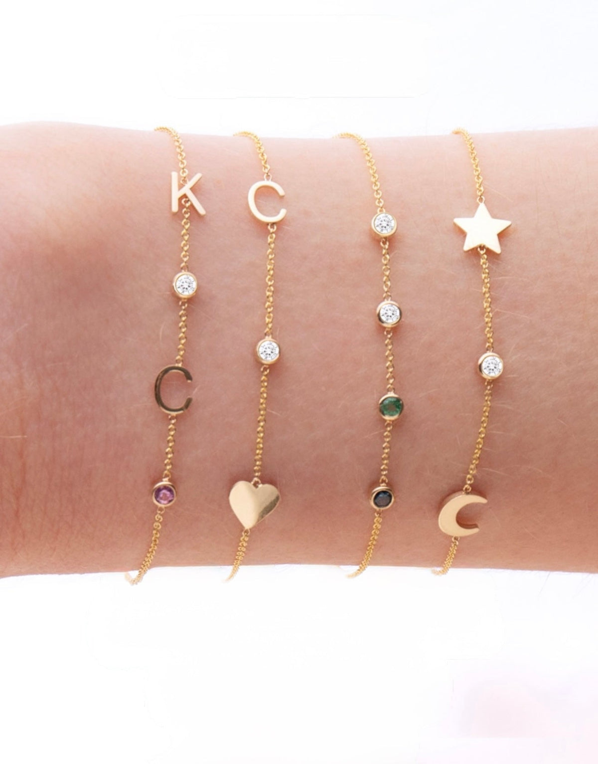 A wrist wearing four yellow gold Custom Gold Bracelets: two with three charms (C, a diamond birthstone and a heart, and a star, a diamond birthstone and a moon) and two with four charms (K, C, diamond and amethyst birthstones, and two diamonds, one emerald and one sapphire), on a white background.