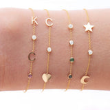 A wrist wearing four yellow gold Custom Gold Bracelets: two with three charms (C, a diamond birthstone and a heart, and a star, a diamond birthstone and a moon) and two with four charms (K, C, diamond and amethyst birthstones, and two diamonds, one emerald and one sapphire), on a white background.