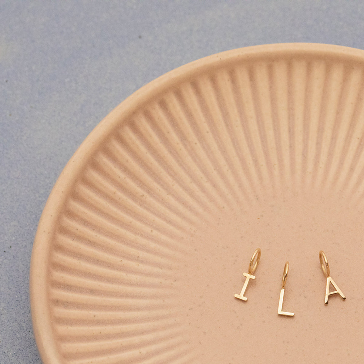 Three Gold Letter Charms in I, L and A on a textured light orange plate with a light grey background. 