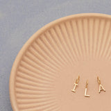 Three Gold Letter Charms in I, L and A on a textured light orange plate with a light grey background. 