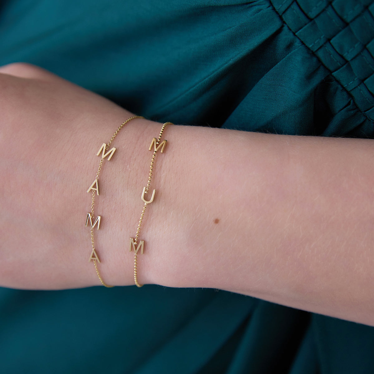 A close-up of a wrist wearing two yellow gold Custom Gold Bracelets in beaded chains, one with four letters M, A, M, A and the other with three letters M, U, M, against a dark turquoise blue silk fabric.